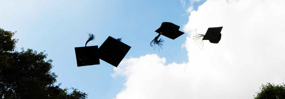 Vier schwarze Doktorhüte fliegen in die Luft vor blauem Himmel mit weißen Wolken.
