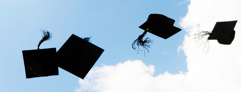 Vier schwarze Doktorhüte fliegen in die Luft vor blauem Himmel mit weißen Wolken.